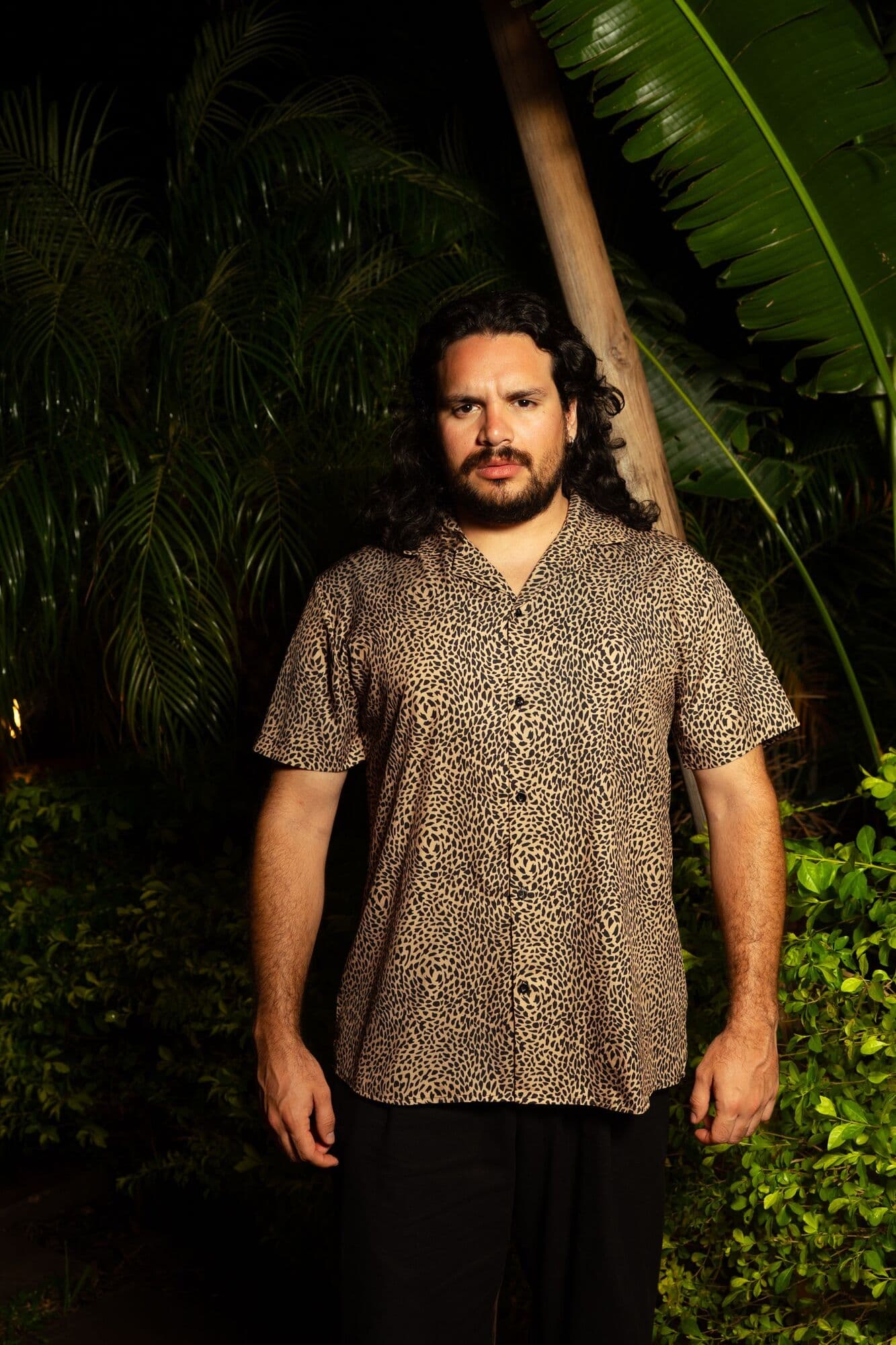 Yisus de frente con camisa de estampado de leopardo entre plantas tropicales de noche
