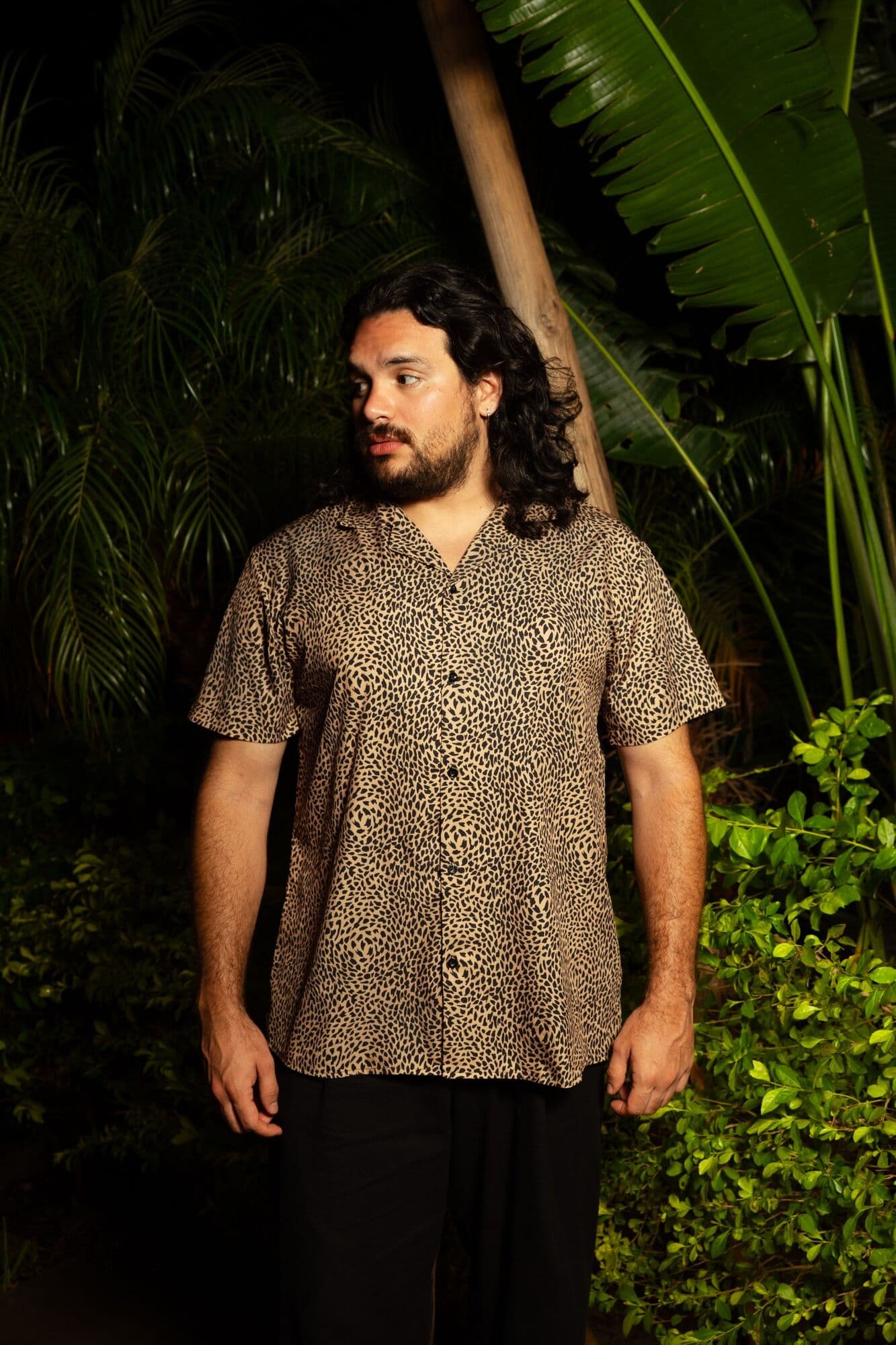 Yisus con camisa de estampado de leopardo, mirando hacia un lado, entre plantas tropicales de noche