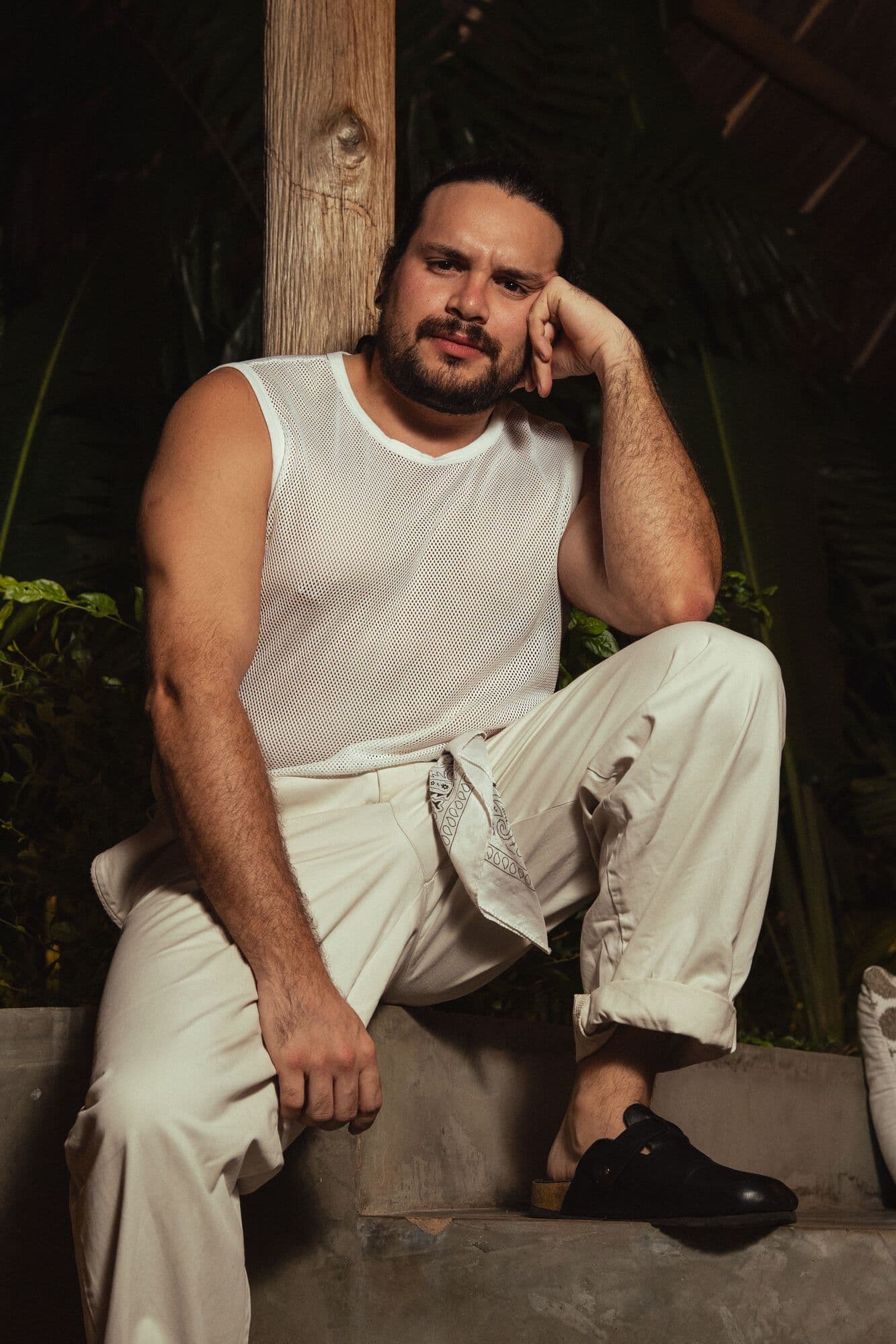 Retrato nocturno de Yisus sentado, con camiseta blanca sin mangas, apoyando la cabeza en la mano